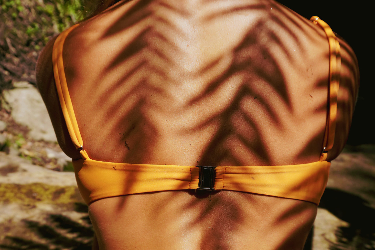 Image of the back of a woman wearing an orange bikini with shadows on her back from the foliage above her.