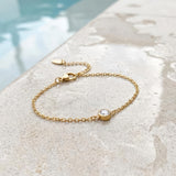 Gold bracelet with a small round gemstone on a textured stone surface on the edge of a pool