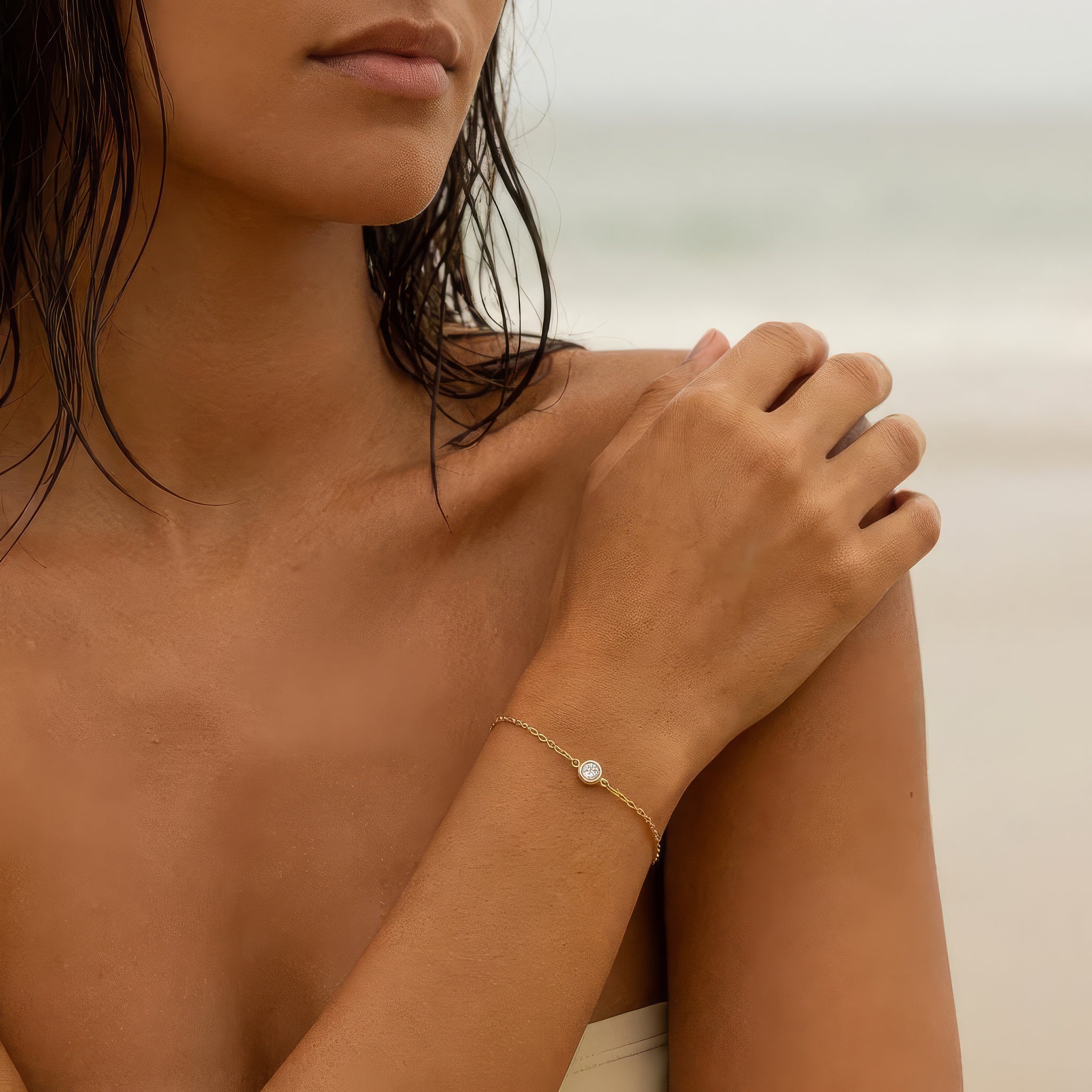 Woman wearing a gold bracelet with a small clear round gemstone on a neutral beach background