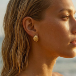 Close-up of a woman wearing gold  shell-shaped earrings with a blurred beach background