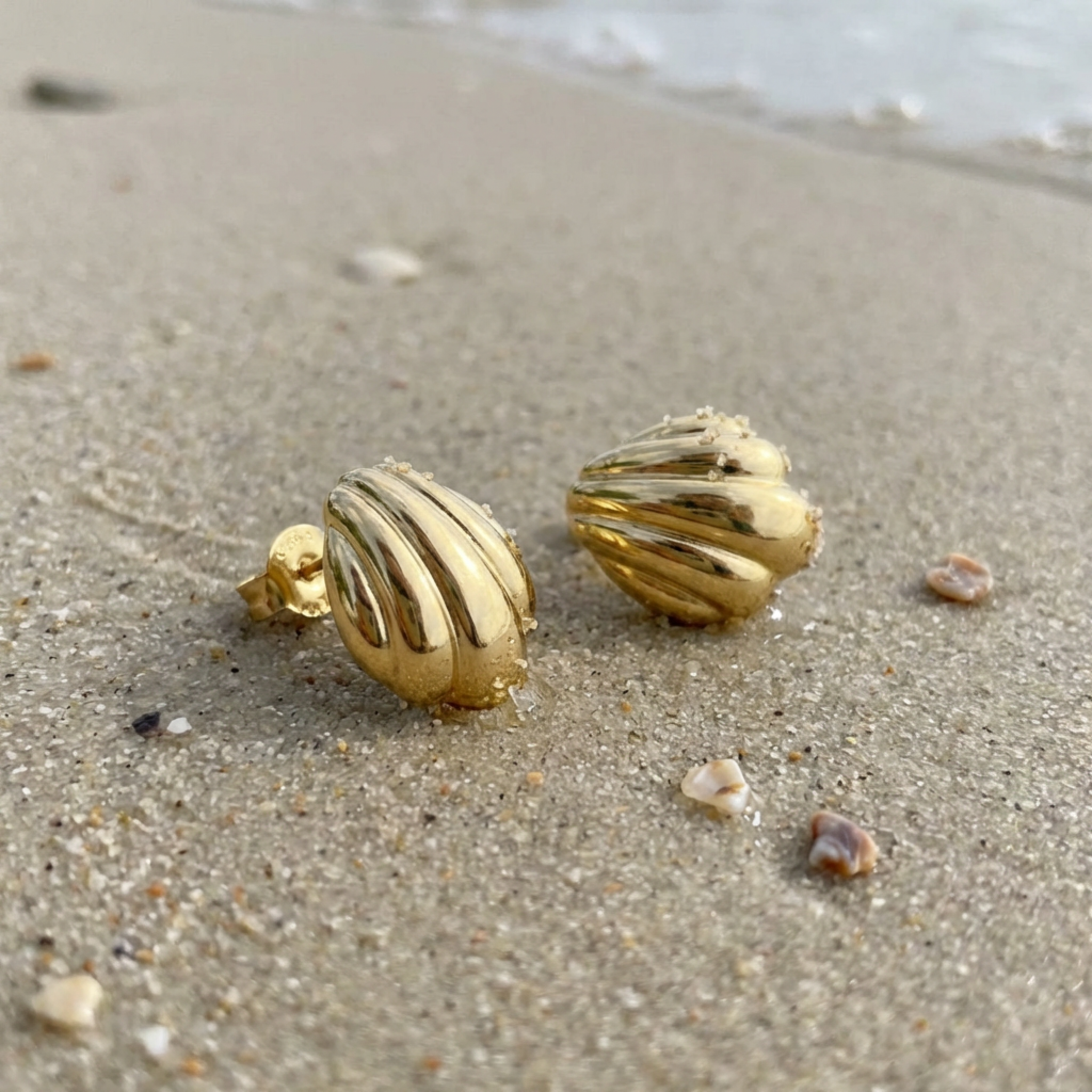 Gold shell-shaped earrings on a sandy beach with the ocean in the background