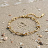 Gold chain bracelet with three diamond-like stones lying in the sand on the beach with the ocean in the background