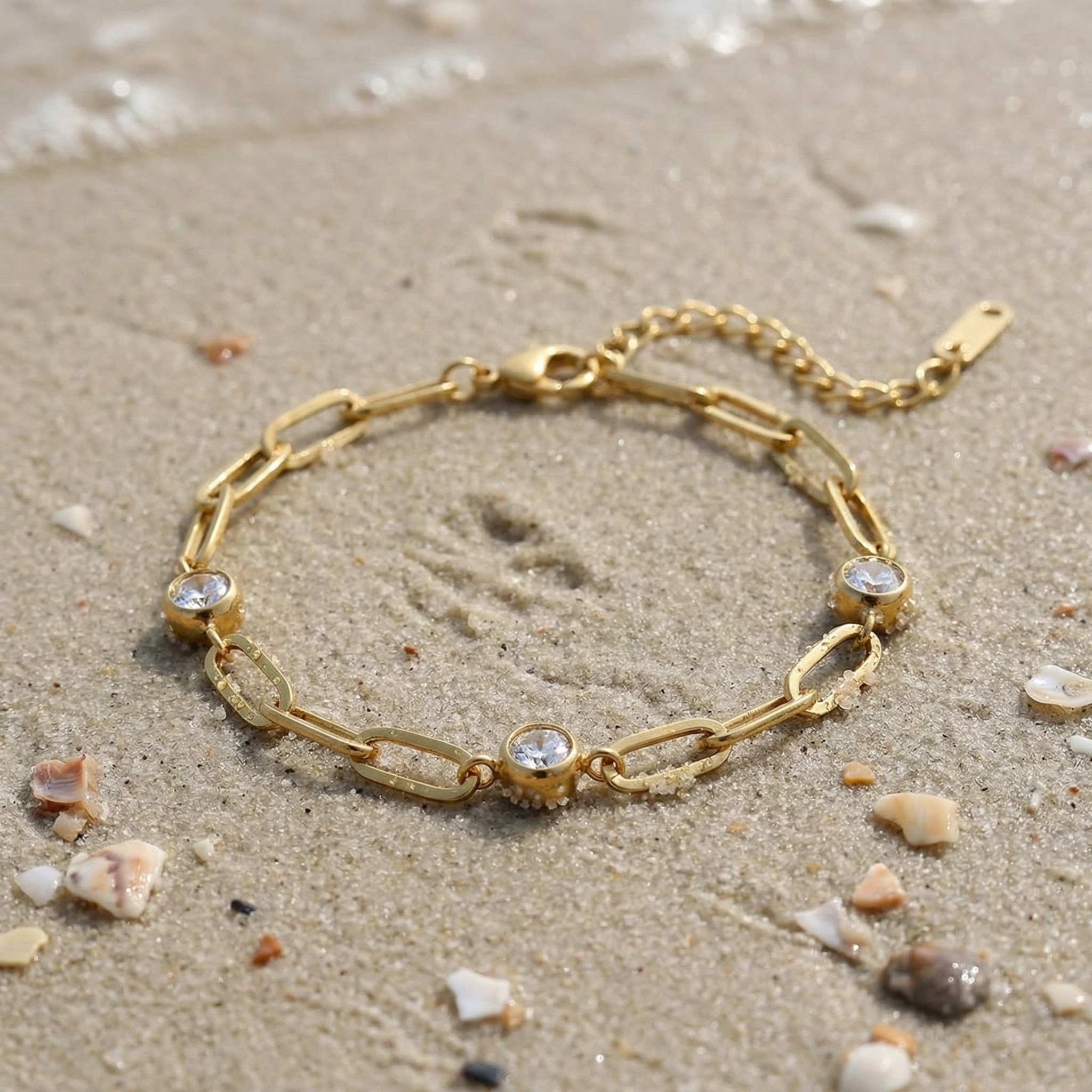 Gold chain bracelet with three diamond-like stones lying in the sand on the beach with the ocean in the background