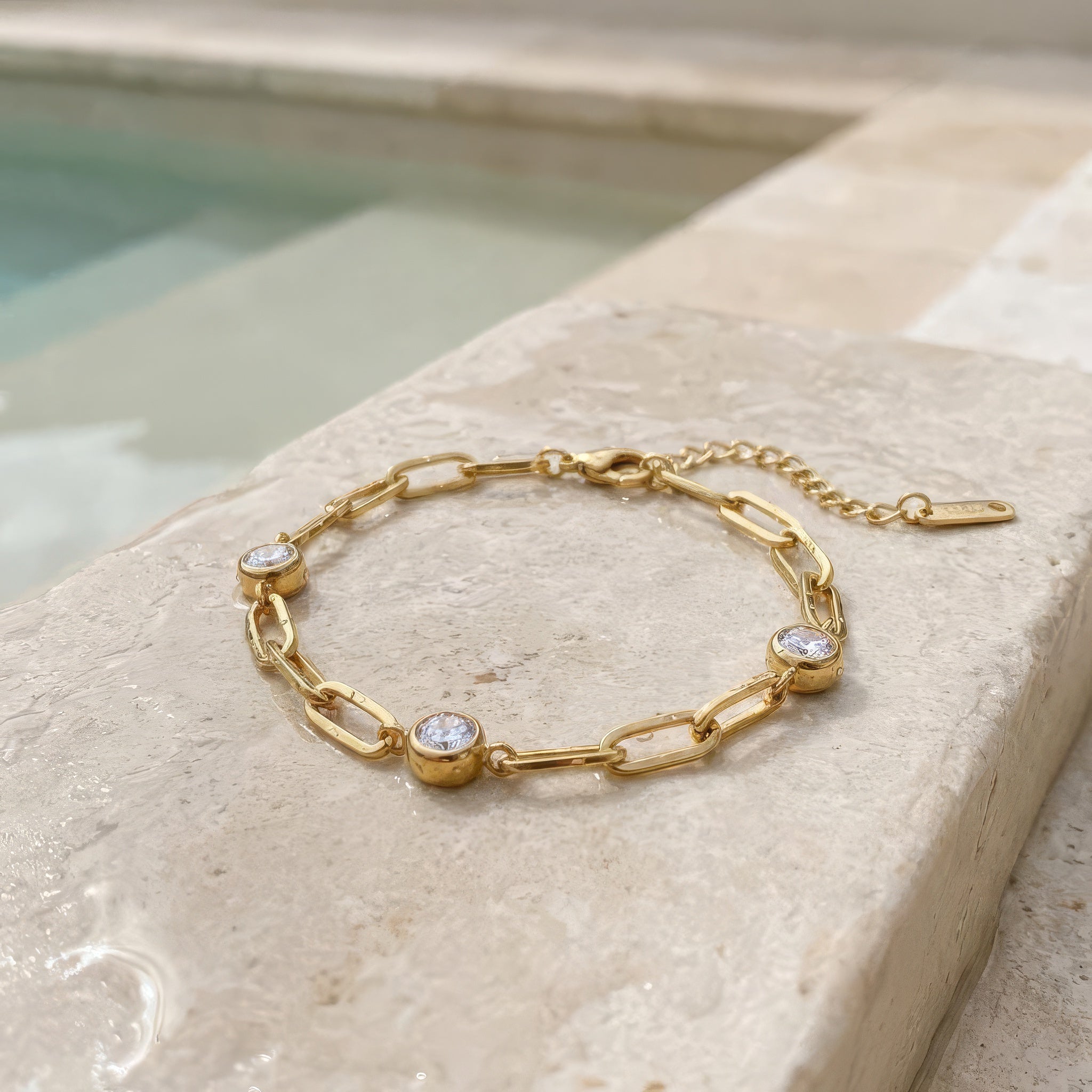 Gold chain bracelet with three diamond-like stones lying on a stone surface on the edge of a pool
