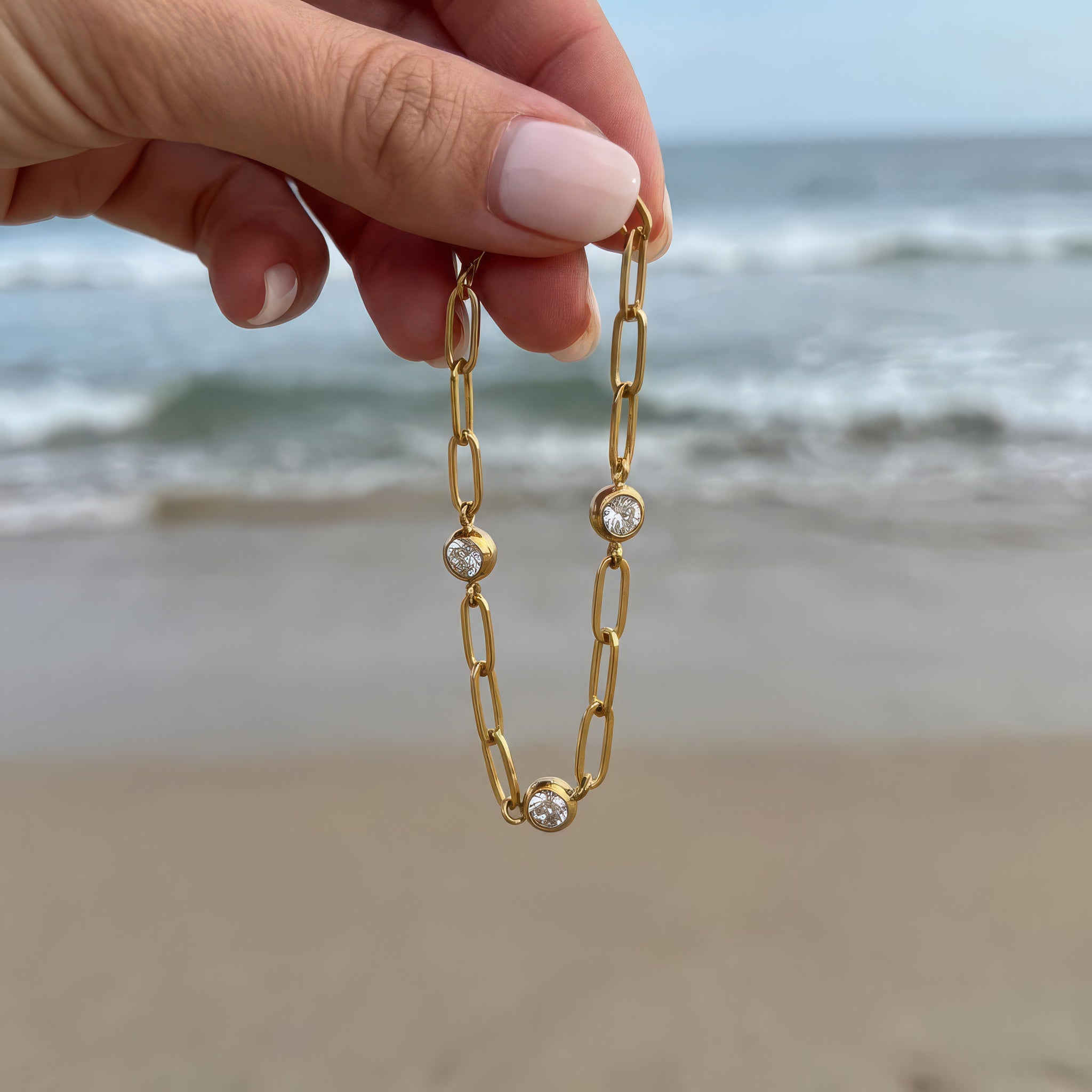 Gold chain bracelet with three diamond-like stones held in hand and dangling in the wind in front of the ocean