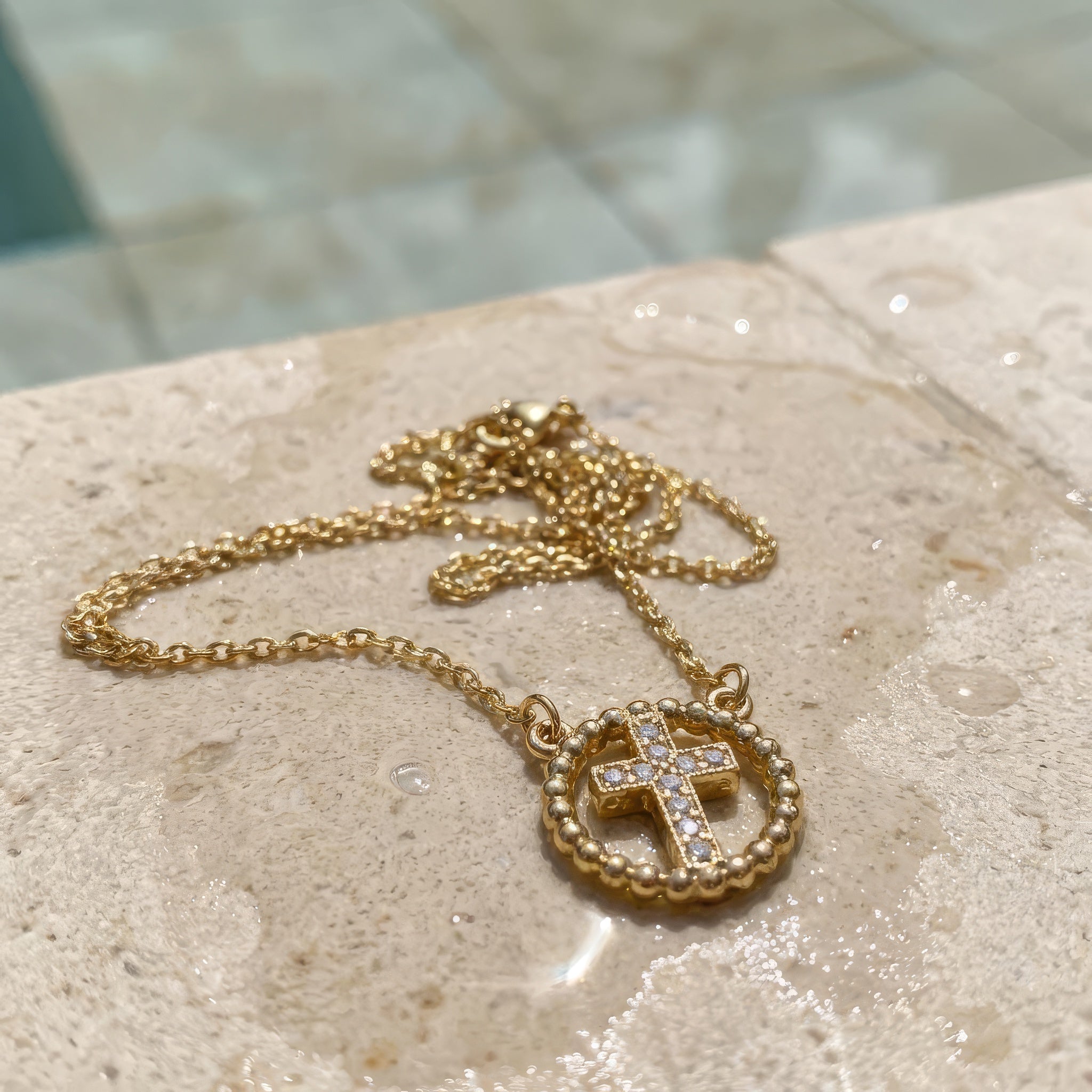 Gold cross necklace with rhinestones lying on the a stone surface at the edge of a pool