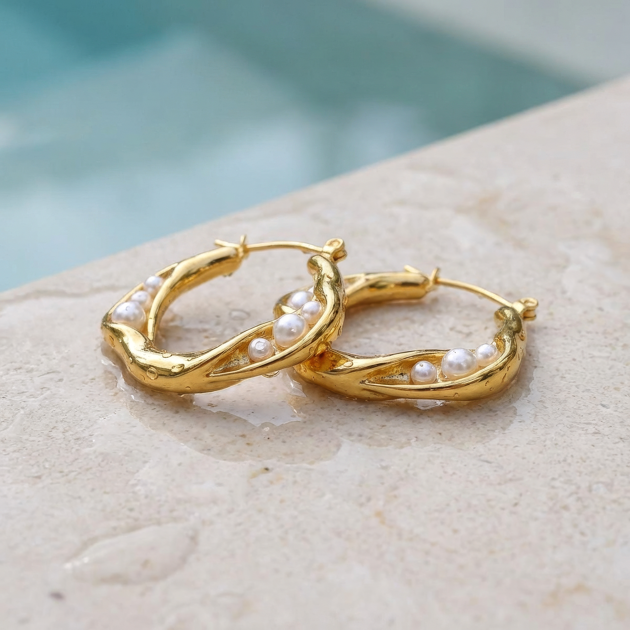 Gold hoop earrings with pearls on a stone surface at the edge of a pool.