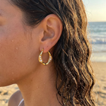 Close-up of a person wearing gold hoop earrings with pearls embedded on a beach background