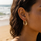 Close-up of a person wearing gold hoop earrings with a beach in the background