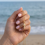 Hand holding a gold cross necklace with ocean in the background