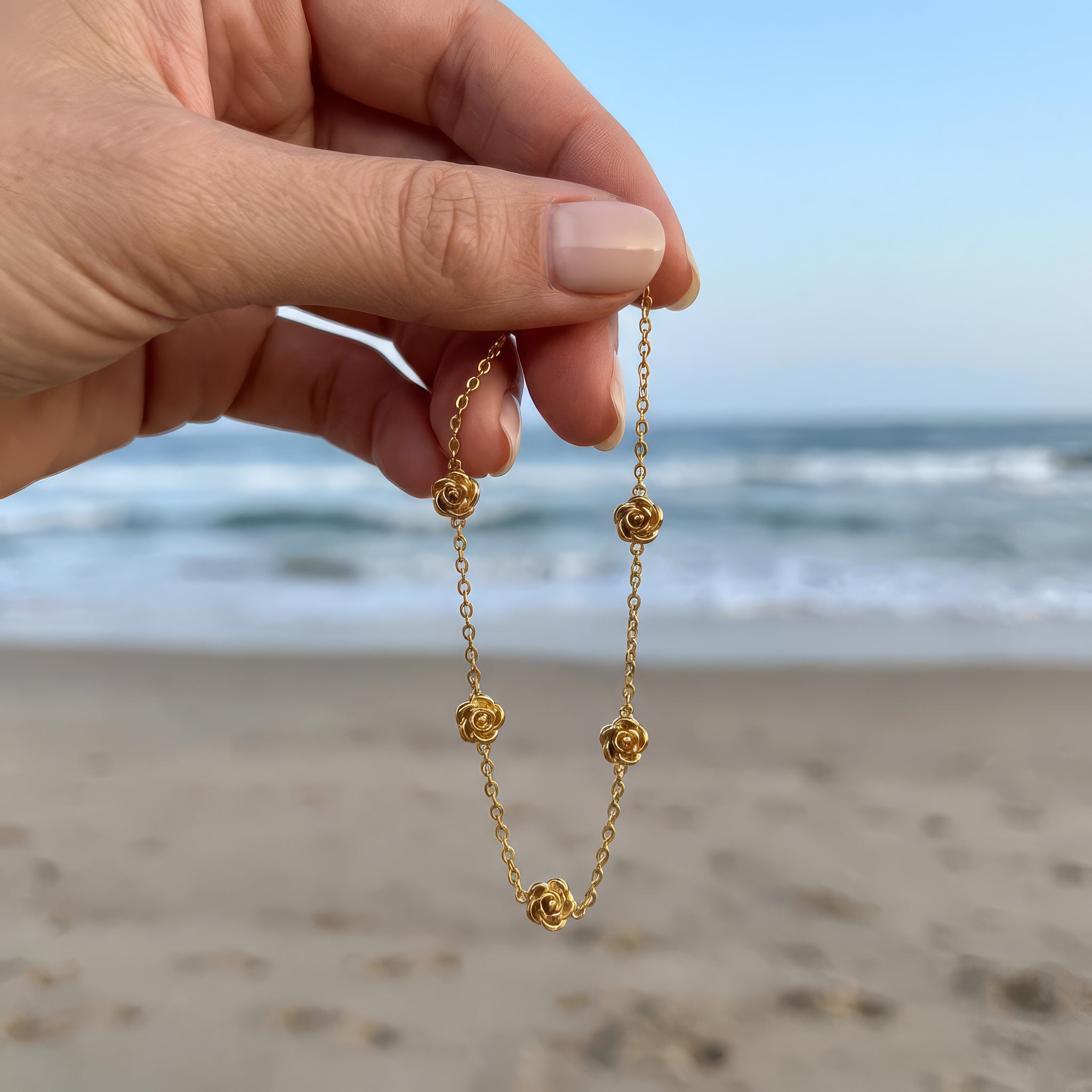 Hand holding a gold bracelet in front of the ocean with five stationary gold rosebud designs spaced out on the chain 