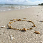 Gold bracelet with five stationary gold rosebud designs spaced out on the chain on a sandy beach with the ocean in the background