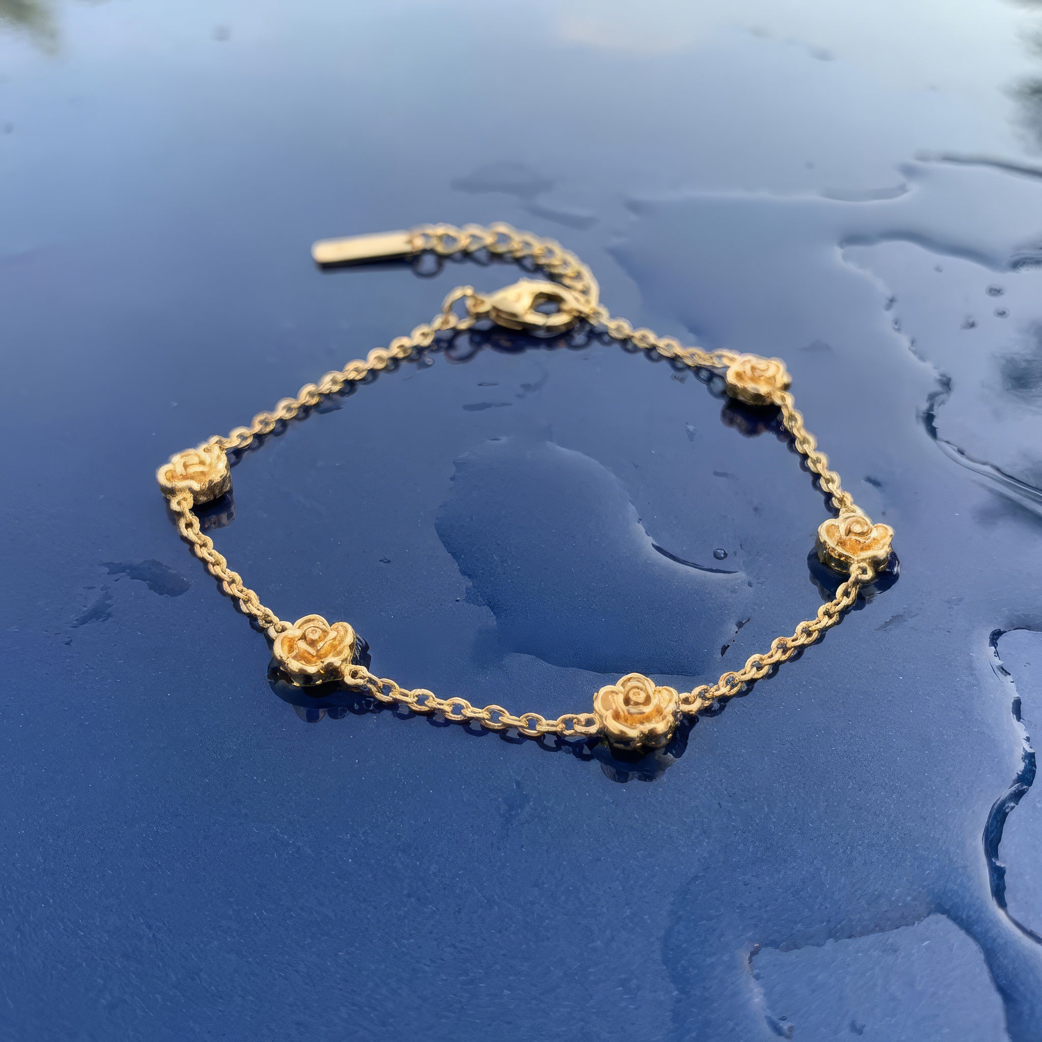 Gold bracelet with five stationary gold rosebud designs spaced out on the chain on a wet blue surface