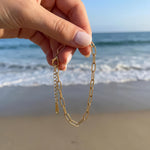 Gold paperclip chain bracelet being held in a womans hand dangling in front of the ocean