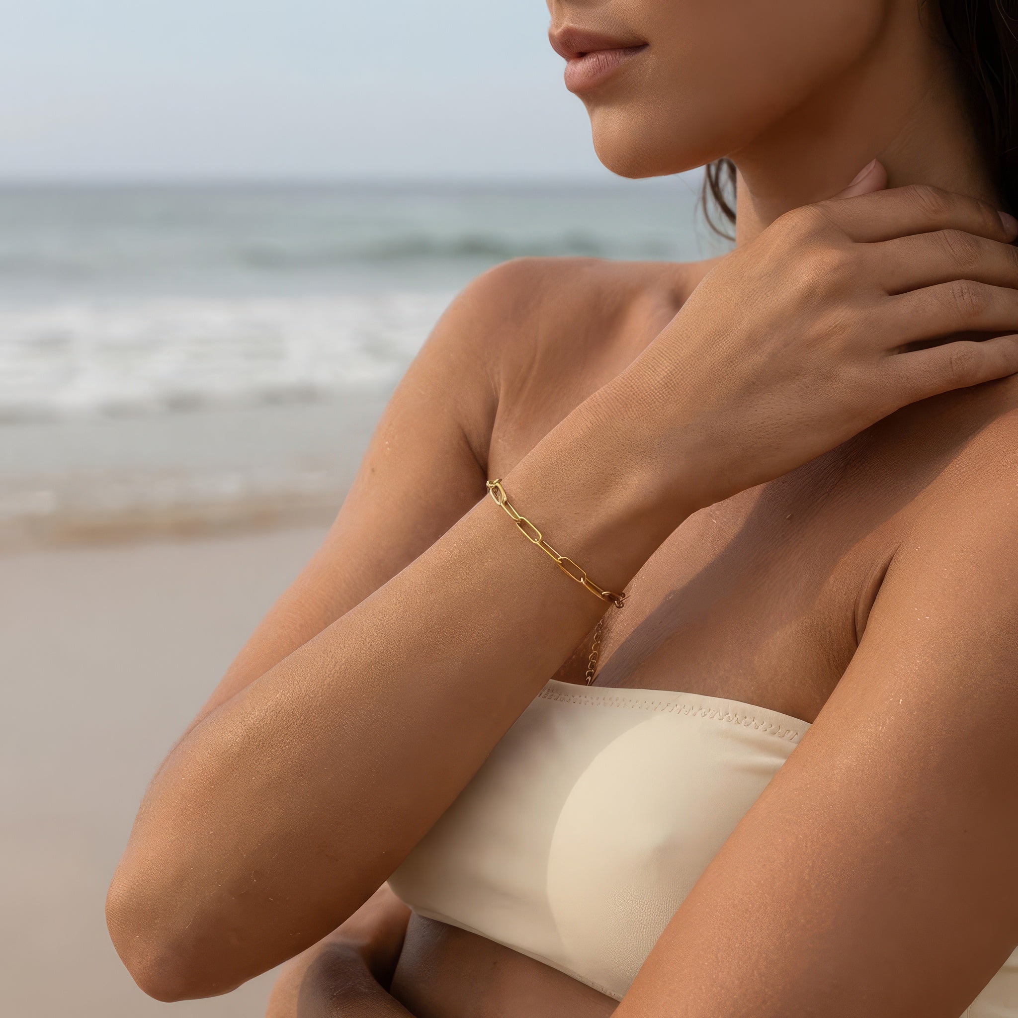 Gold paperclip chain bracelet on a woman wearing an off white bathing suit at the ocean