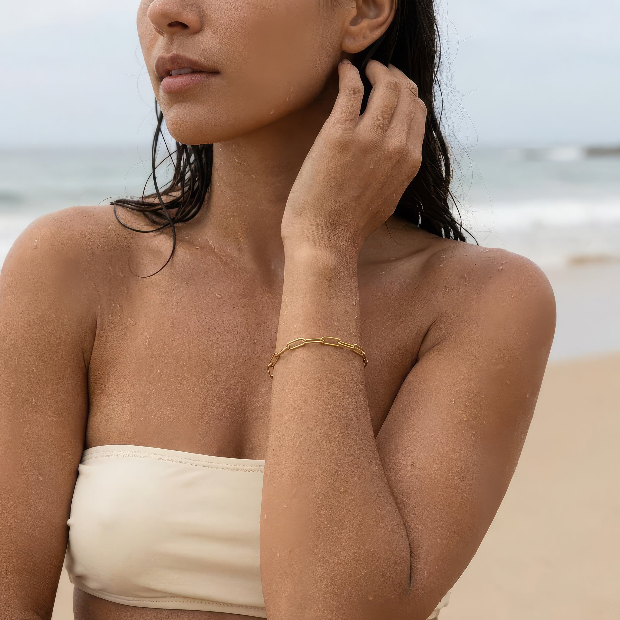 Gold paperclip chain bracelet on a woman wearing an off white bathing suit at the ocean