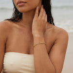 Gold paperclip chain bracelet on a woman wearing an off white bathing suit at the ocean