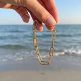 Hand holding gold paperclip chain bracelet dangling in front of the ocean